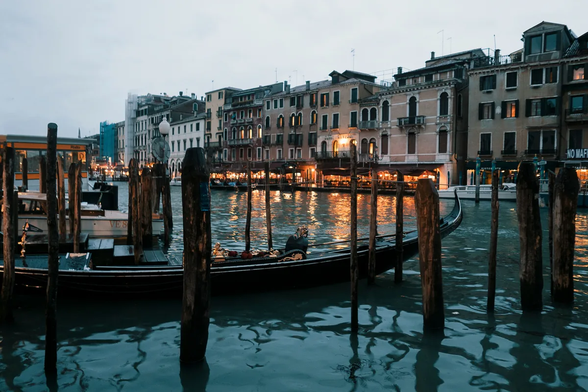 A gondola moored on a Venice canal at dusk with lit palazzo windows reflected in the water, representative of Venice after dark