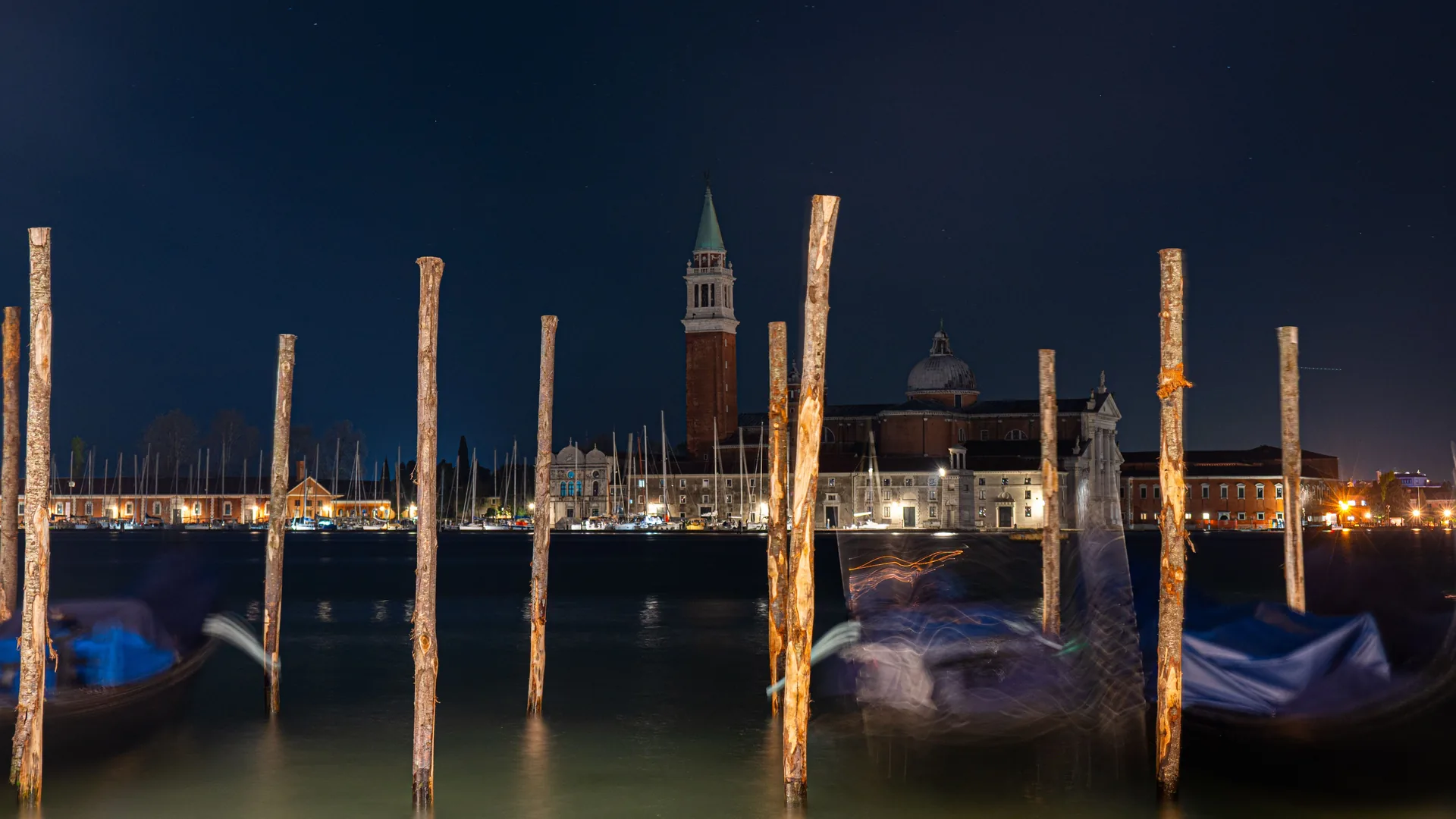 A dark lagoon at night with mooring poles and a church bell tower on the far shore, representative of the view near San Giorgio Maggiore